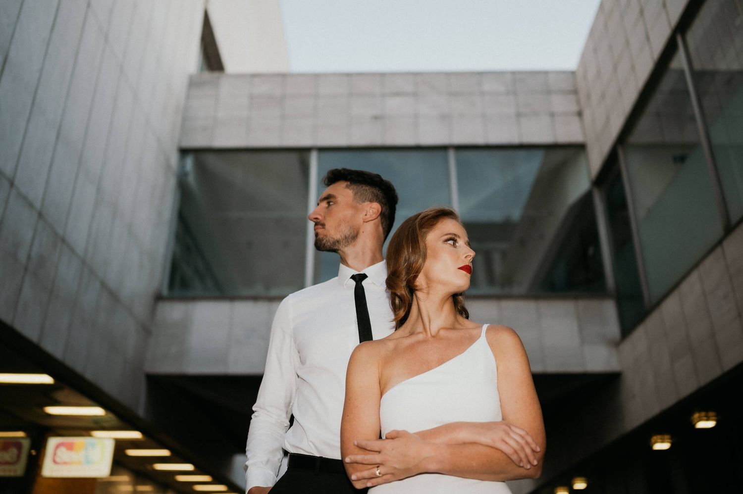 A stylish couple in white and black attire poses dramatically against a modern architectural backdrop.