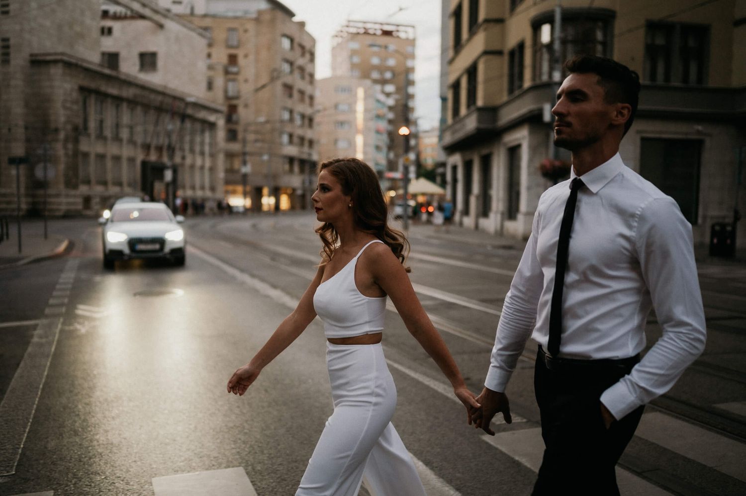 A couple in matching white and black outfits walk playfully through city streets at dusk.