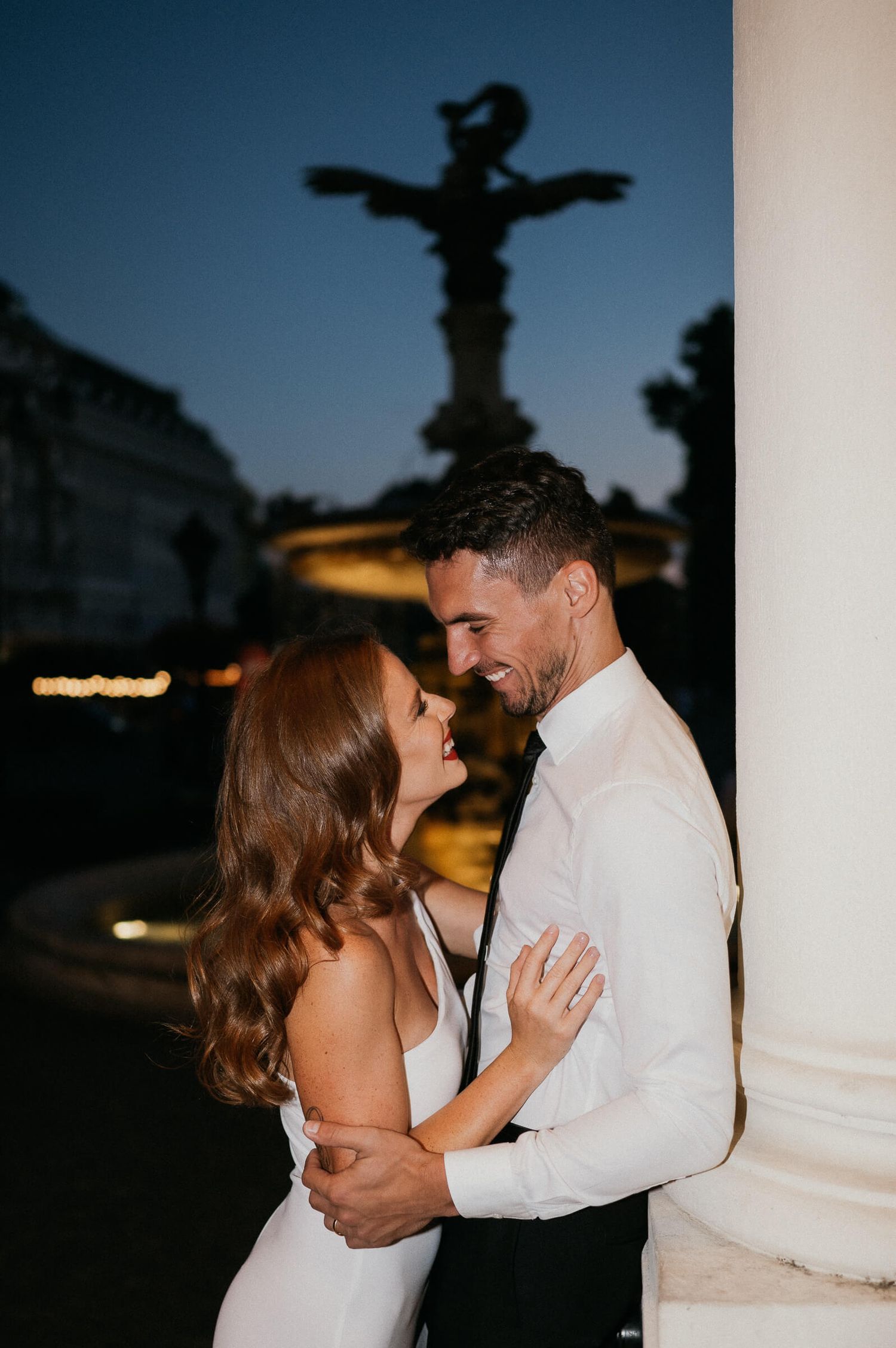 A romantic couple embraces at twilight with an ornate statue silhouetted in the background.