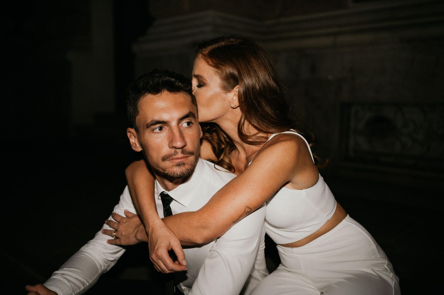 A couple in formal attire shares an intimate moment in a darkened setting.