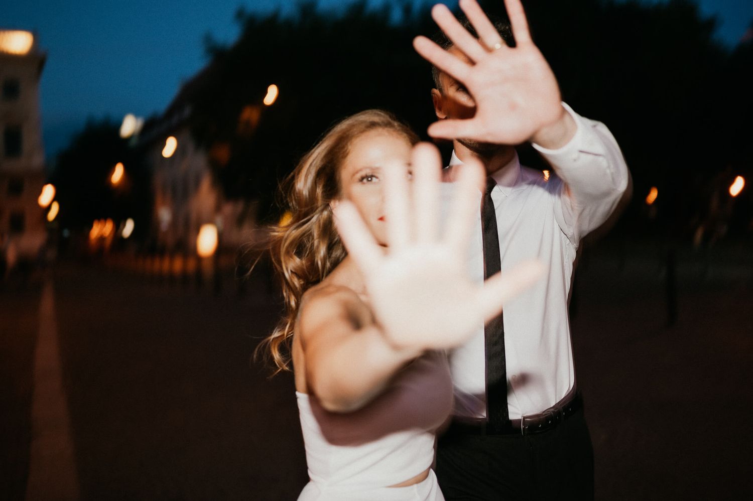 A playful couple in wedding attire raises their hands toward the camera in an evening setting.