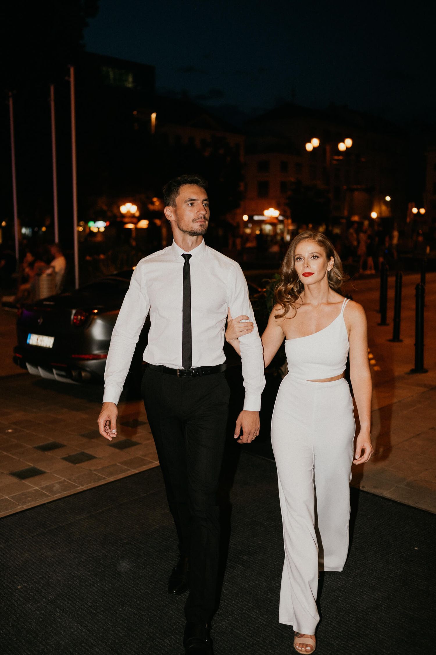 A couple in matching white and black formal wear walks together on a city street at night.