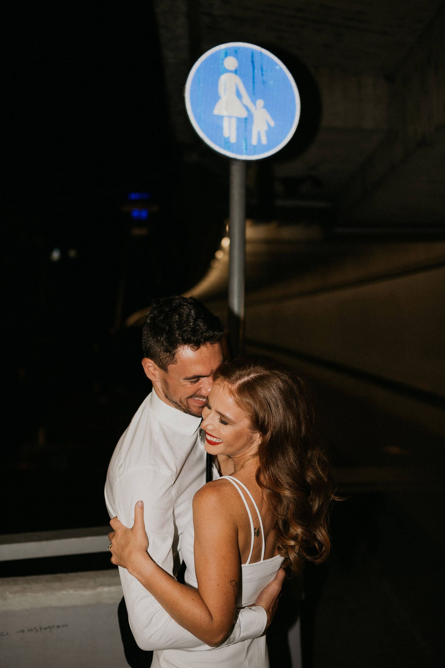 A couple embraces beneath a pedestrian crossing sign during a nighttime photoshoot.
