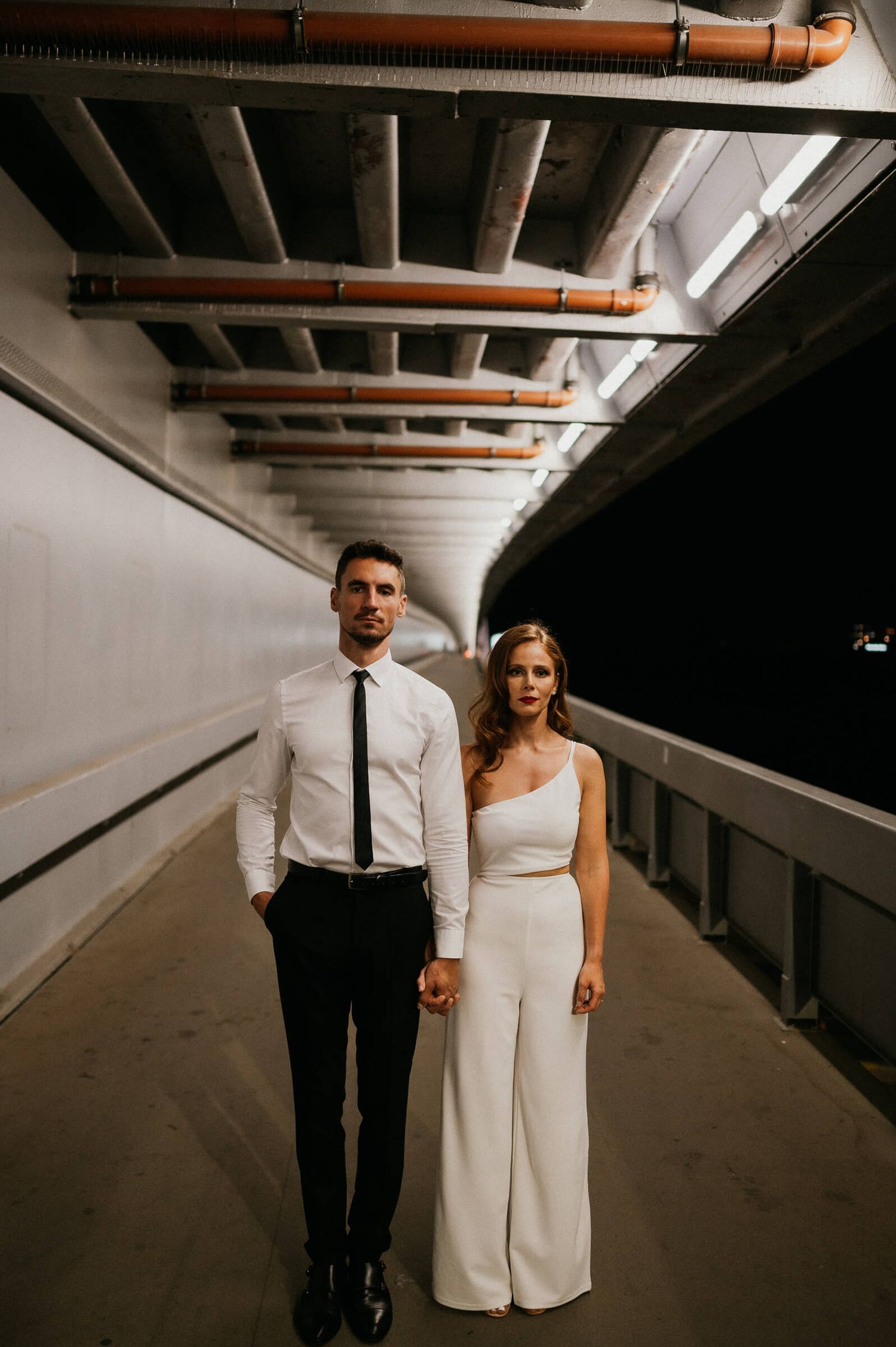 A couple in formal wear poses in an industrial corridor with exposed ceiling pipes.