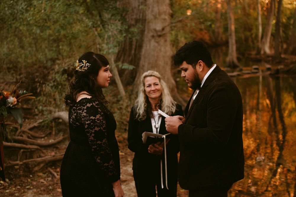 Outdoor wedding ceremony in a wooded area with couple and officiant exchanging vows.