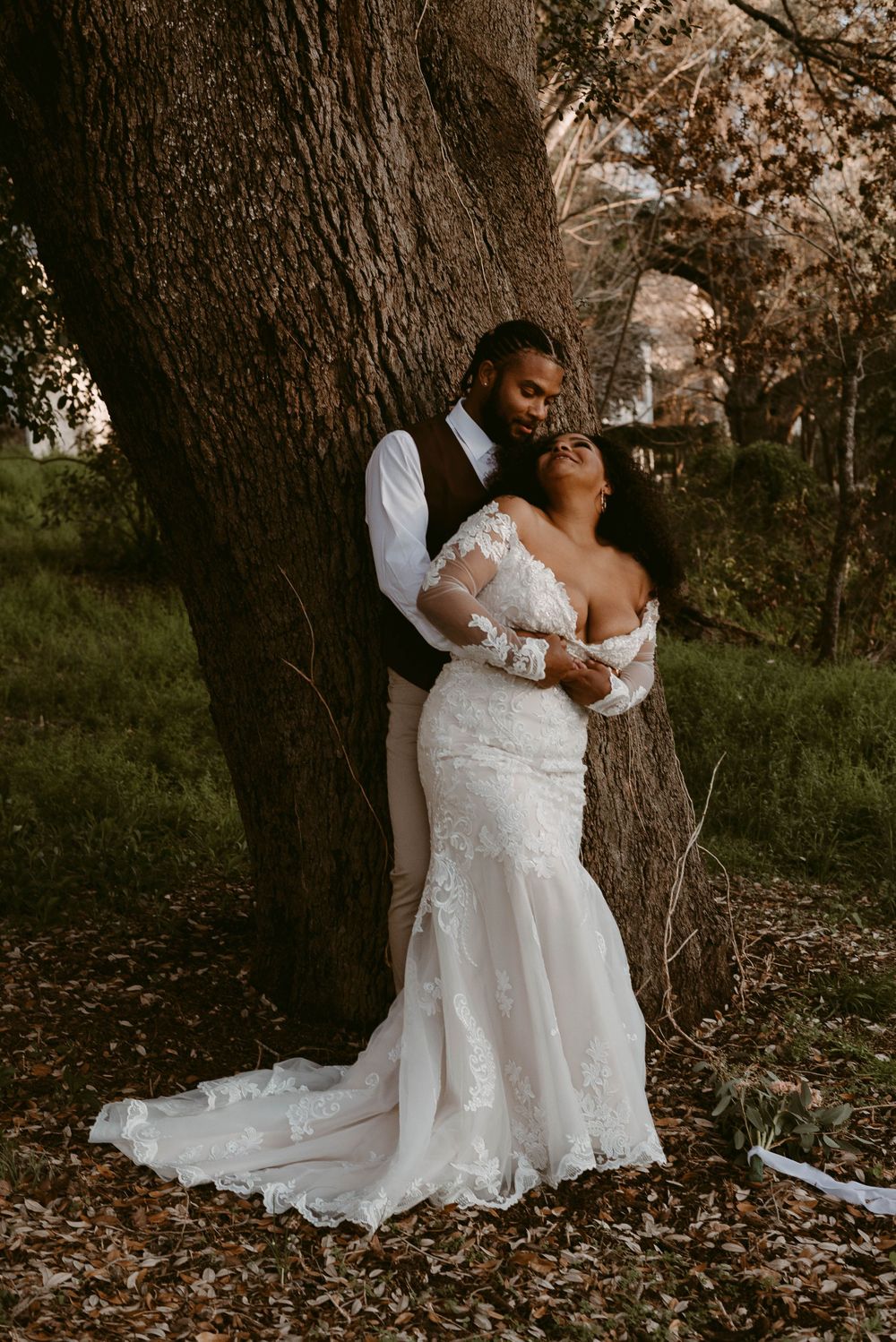 A romantic wedding portrait of a couple in white attire embracing against a rustic tree backdrop.