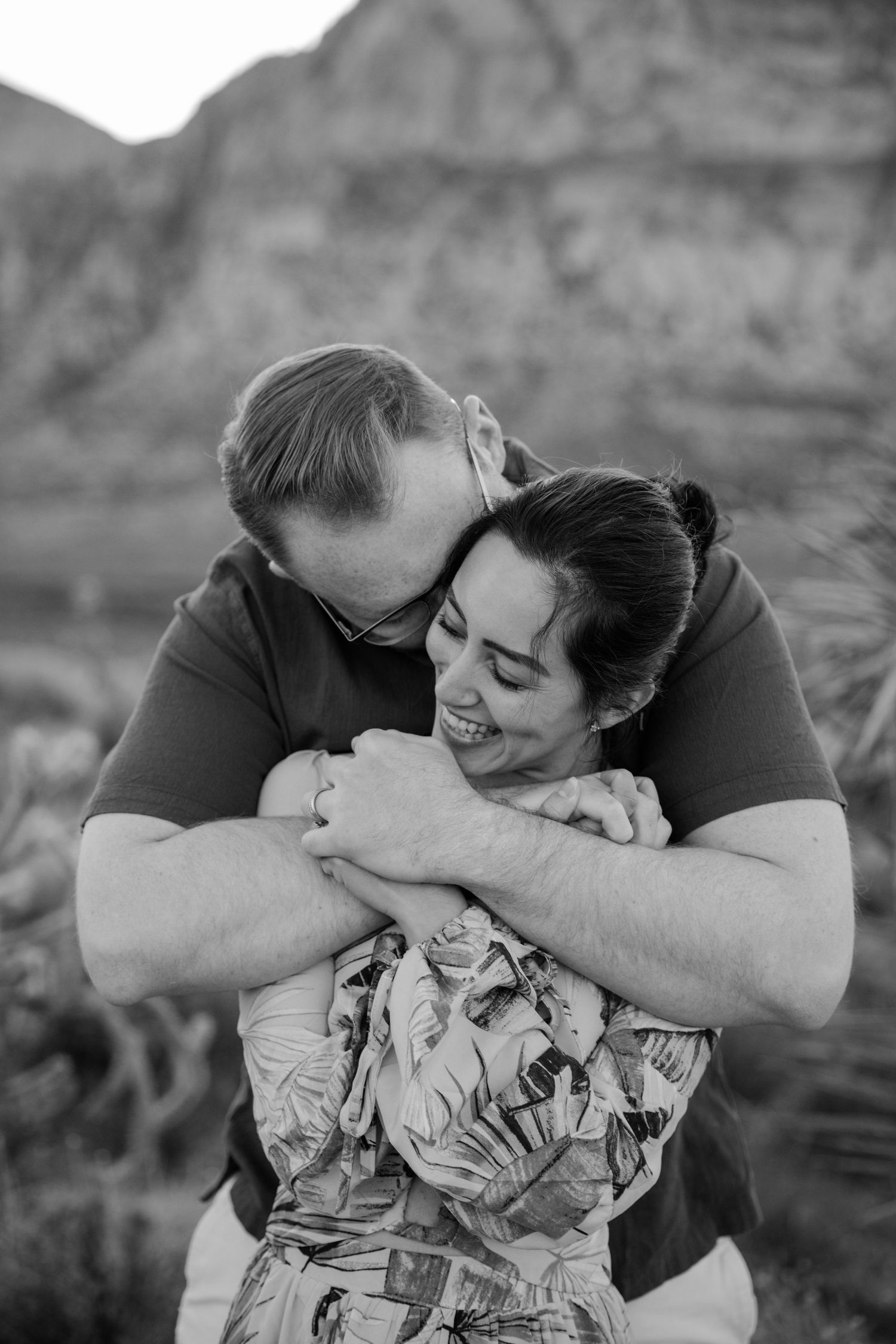 Intimate black and white portrait of couple embracing against desert mountain backdrop.