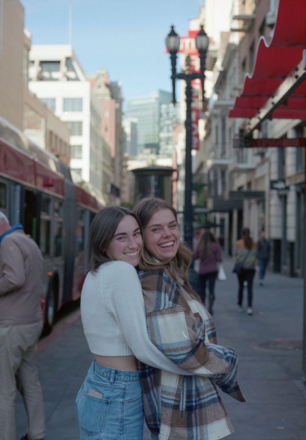 Two friends sharing a laugh on a city street with vintage lamp posts and storefronts in the background.