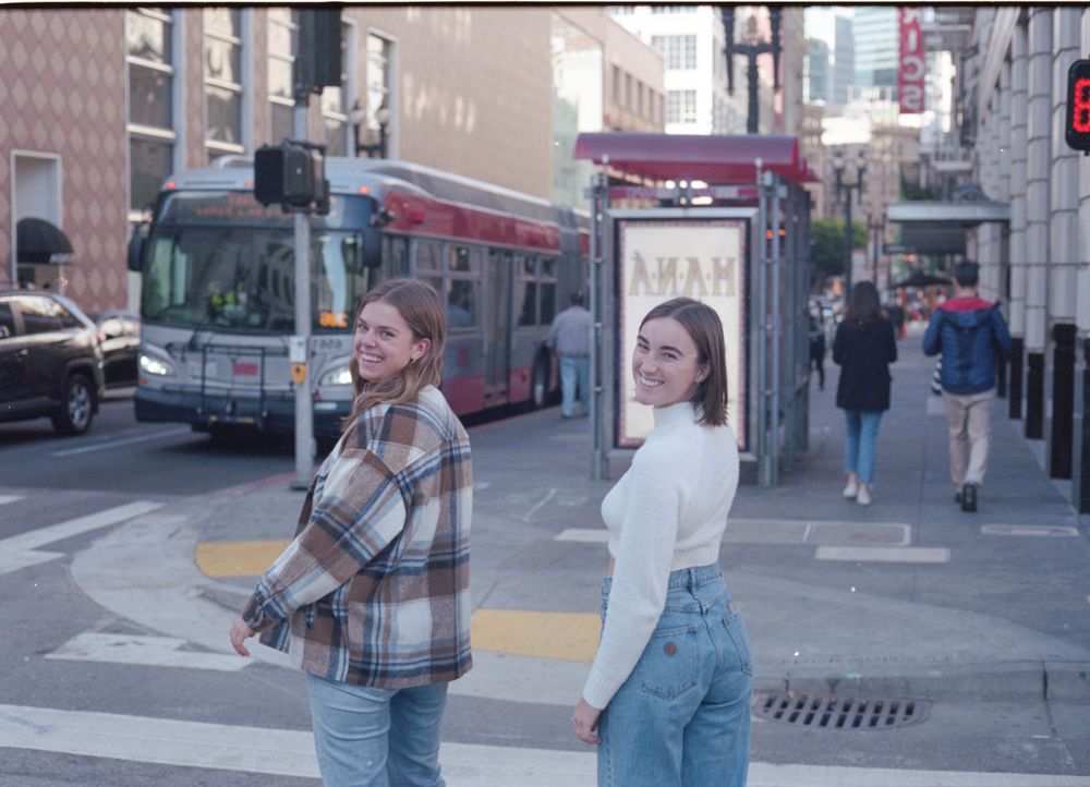 Casual friends posing on an urban street corner with a city bus visible behind them.