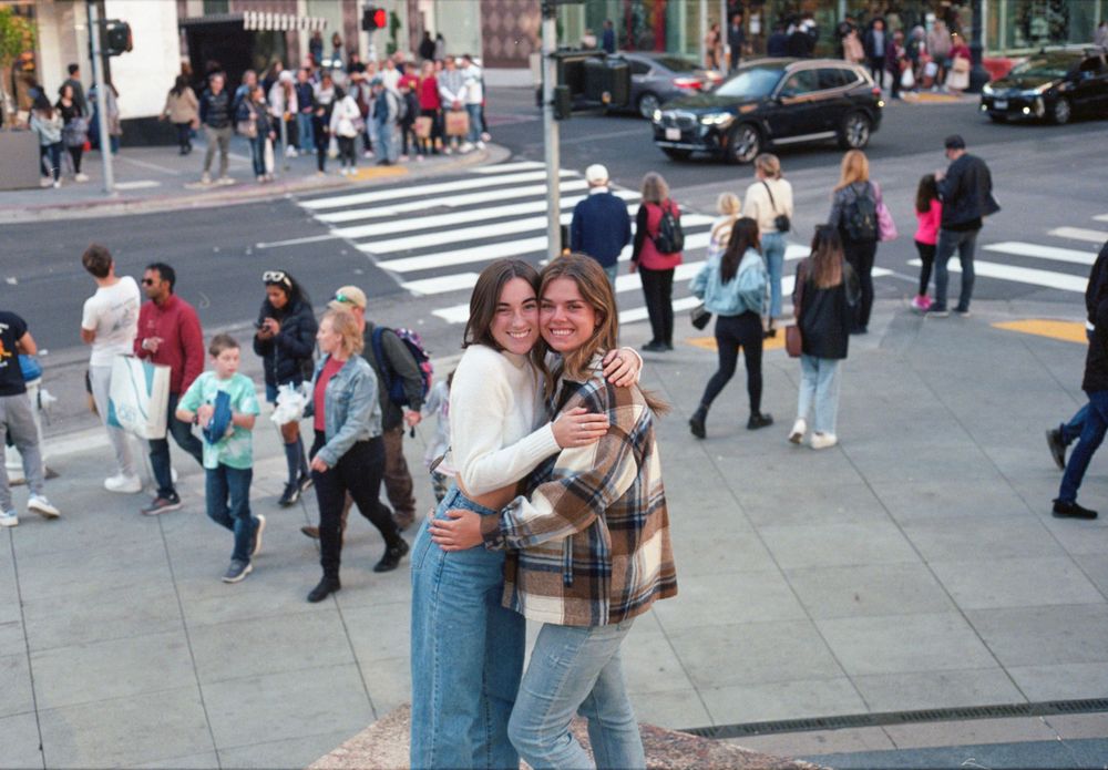 Friends embracing on a busy city street corner with pedestrians walking by.