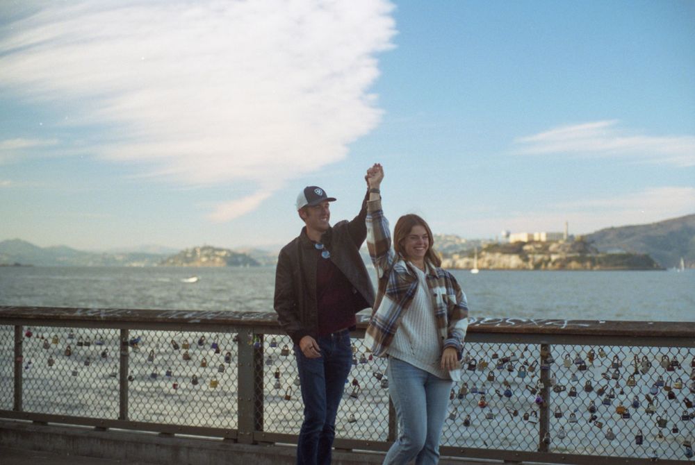 Couple celebrating with raised arms against San Francisco Bay backdrop with Alcatraz visible.