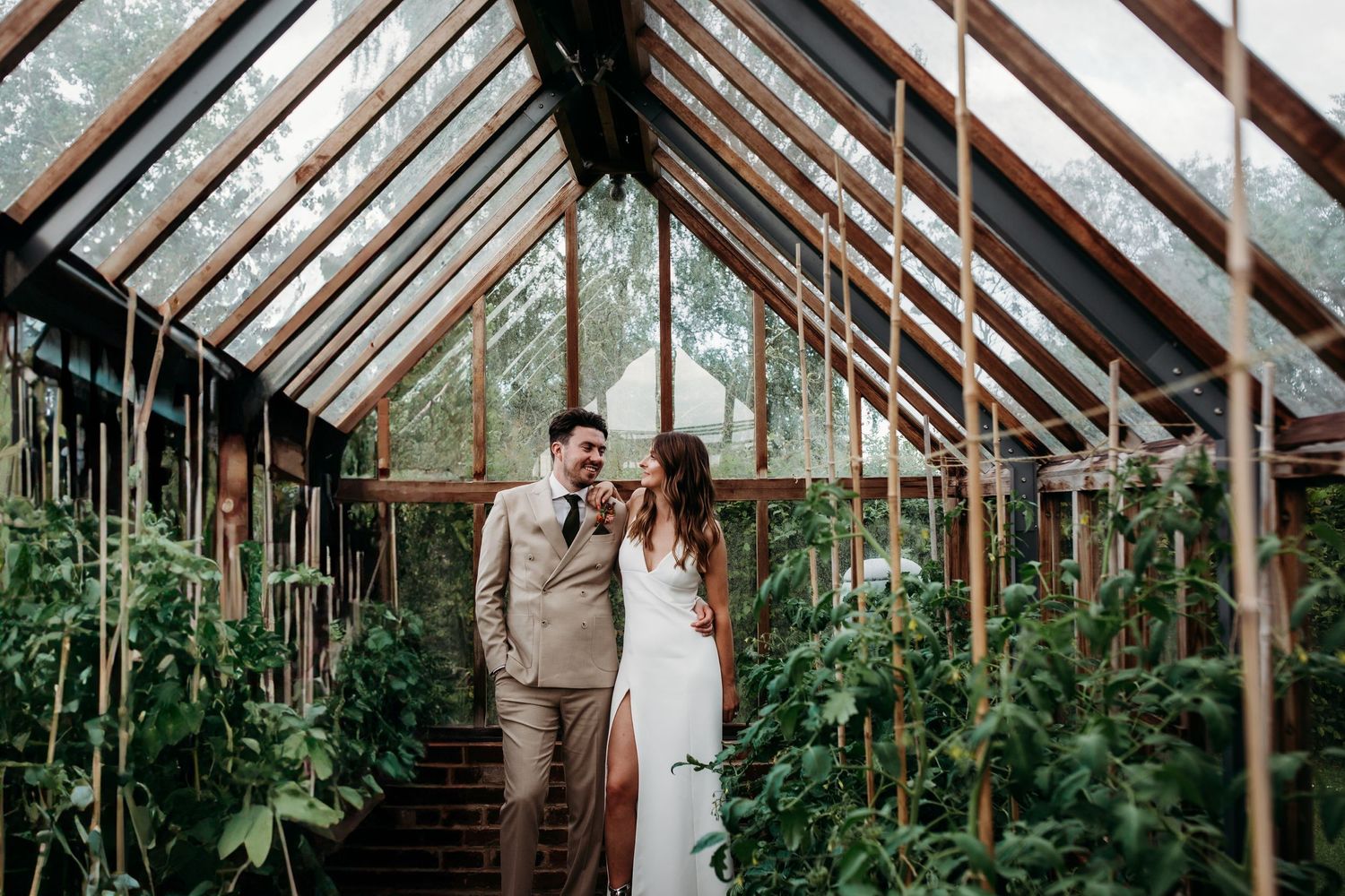 A couple poses inside a vintage greenhouse filled with climbing plants and tomato vines.
