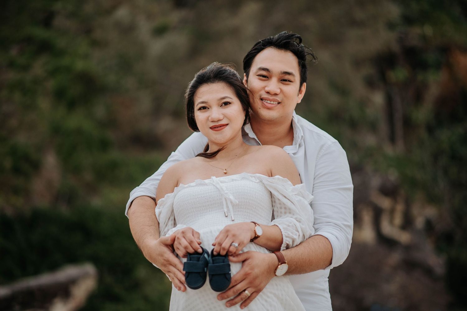 A couple holding baby shoes in a tender embrace outdoors.