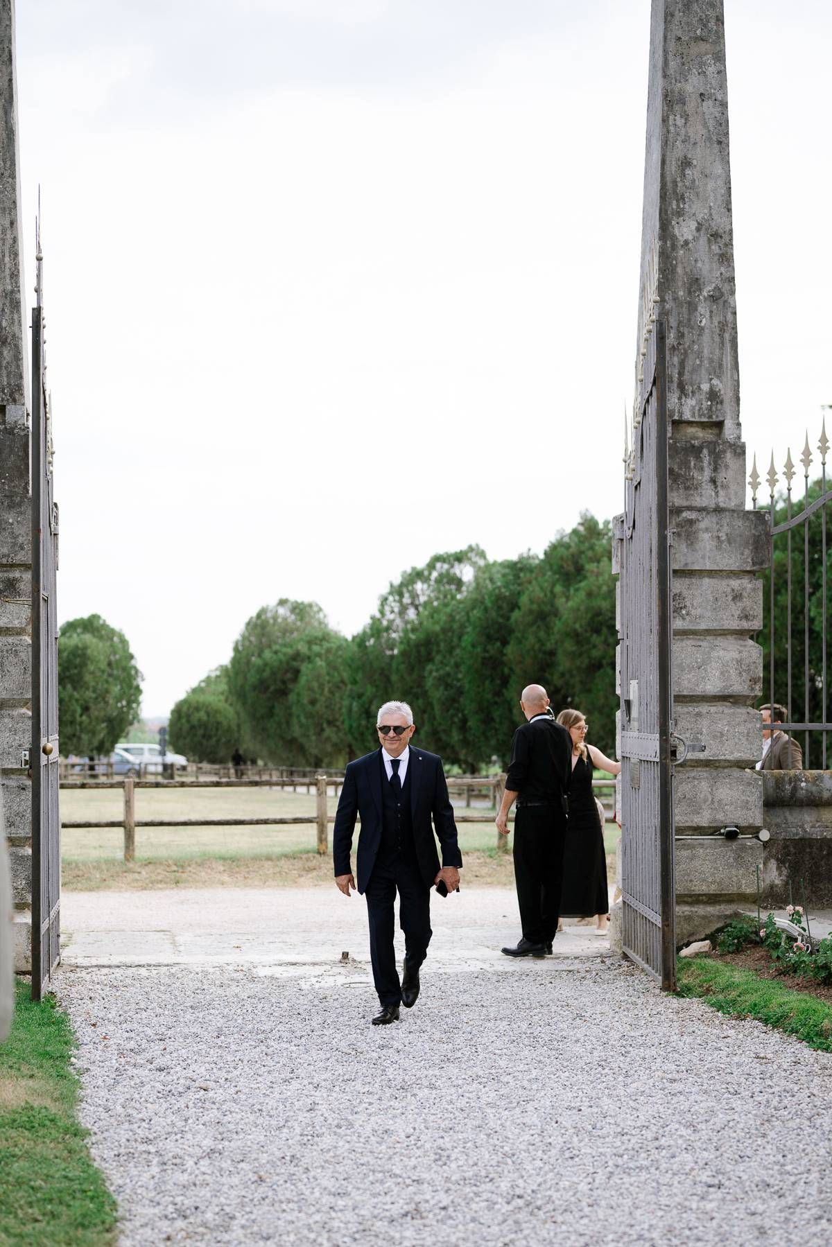 Person in dark suit walking through grand stone gate entrance on gravel path.