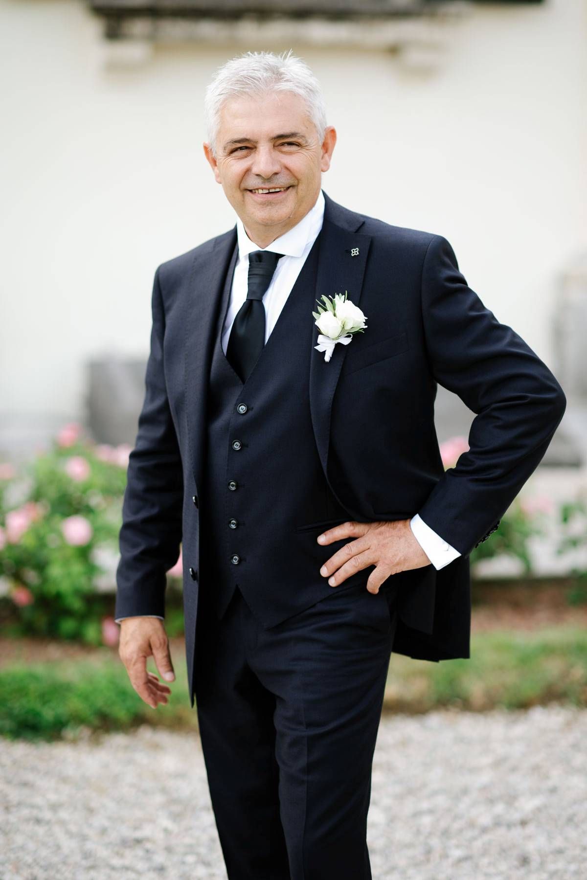 Distinguished gentleman in formal navy suit with white boutonniere smiling at camera.