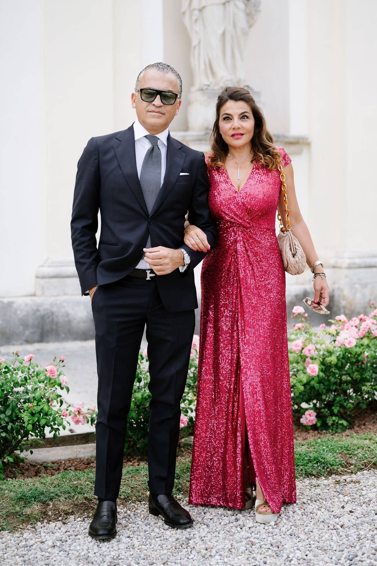 Elegant couple in formal attire posing against white classical architecture with garden.