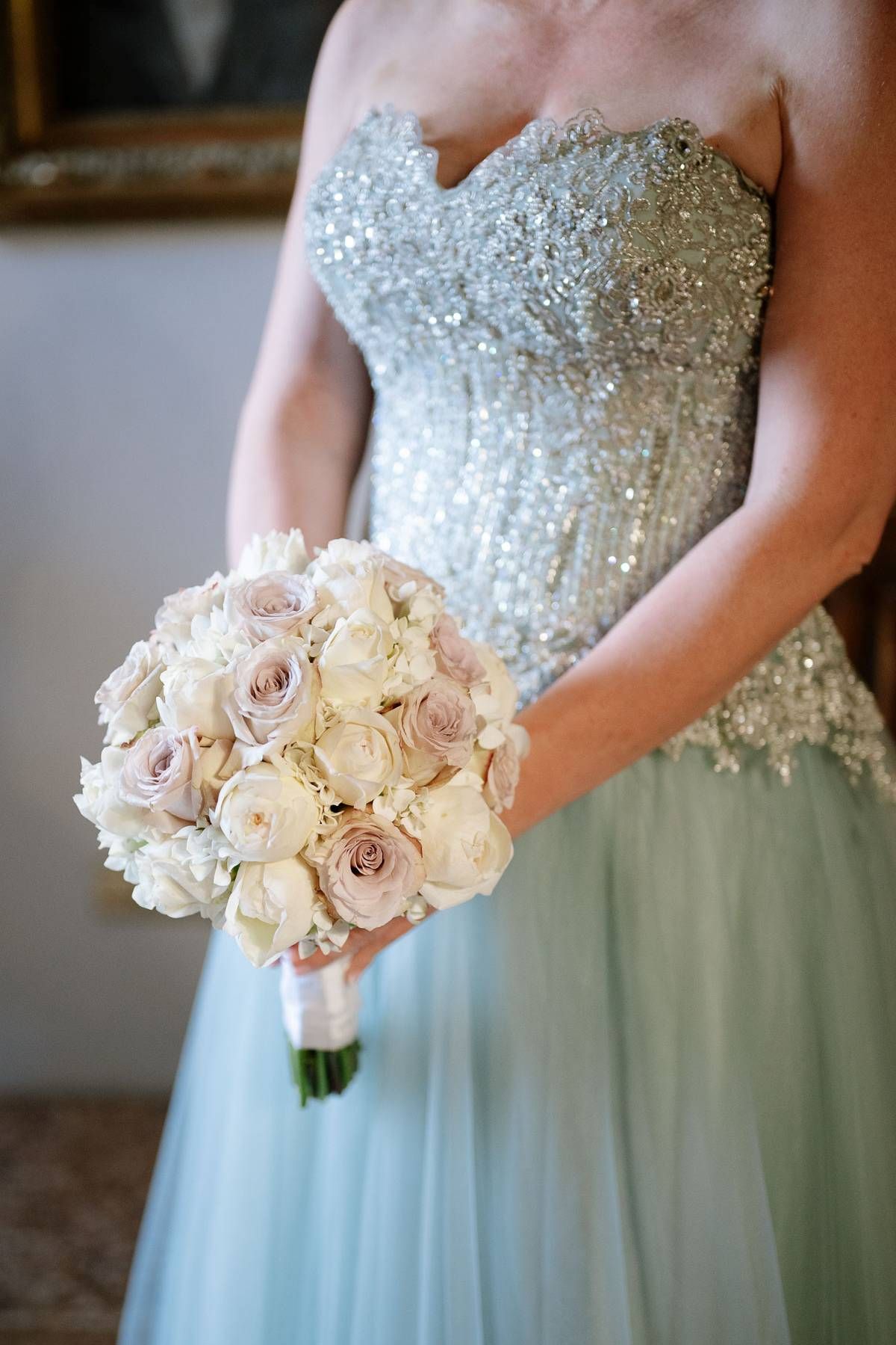 An elegant beaded wedding dress bodice is shown with a soft blue tulle skirt and delicate cream and blush rose bouquet.