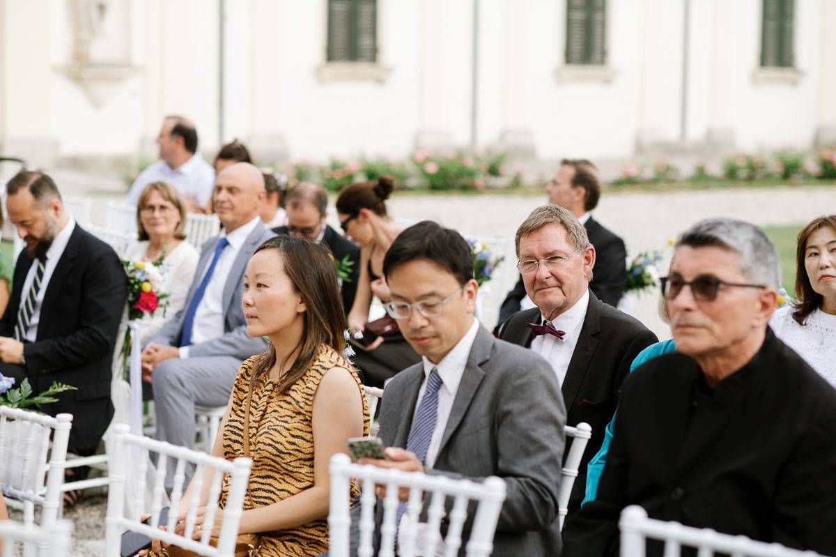 Wedding guests seated in white chairs watch the ceremony during an outdoor celebration.