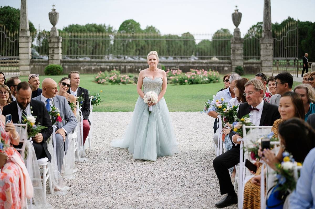 A formal outdoor wedding ceremony with guests seated on white chairs along a gravel path surrounded by manicured gardens.