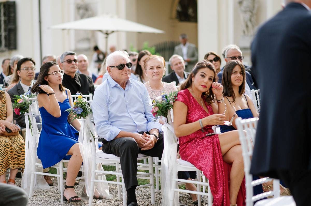 Wedding guests in colorful attire seated on white chairs at an outdoor ceremony.