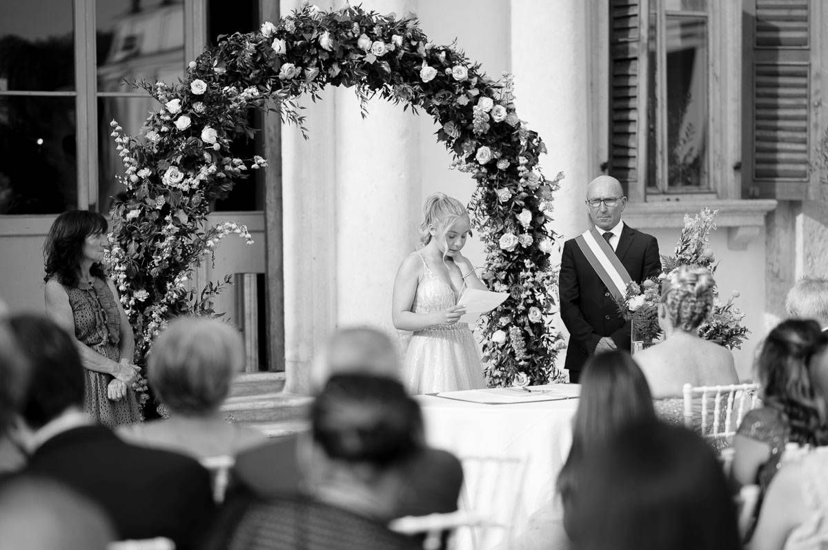 A black and white photograph of a wedding ceremony under a floral arch with guests watching.
