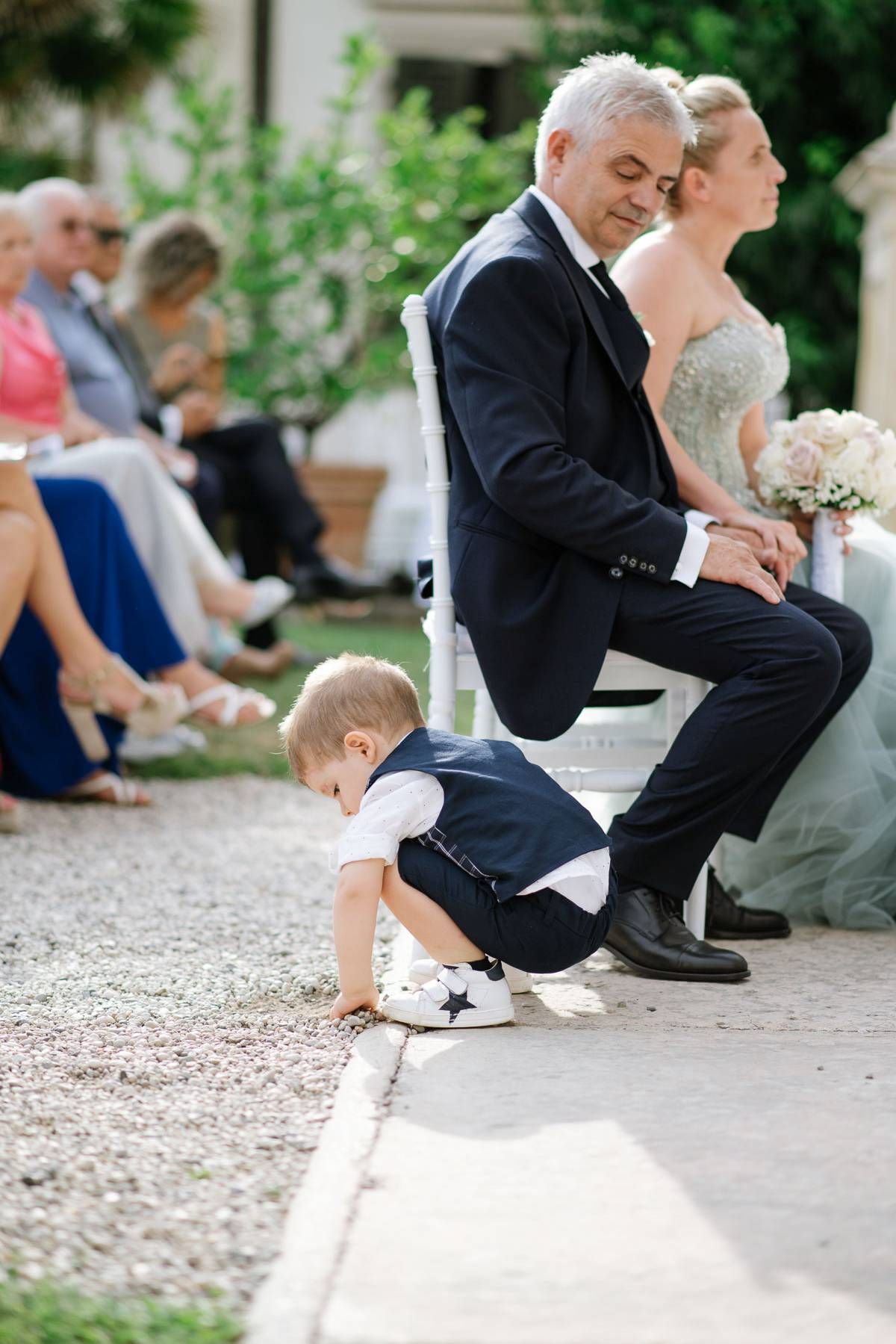 Young child in formal attire plays near guests during outdoor wedding reception.