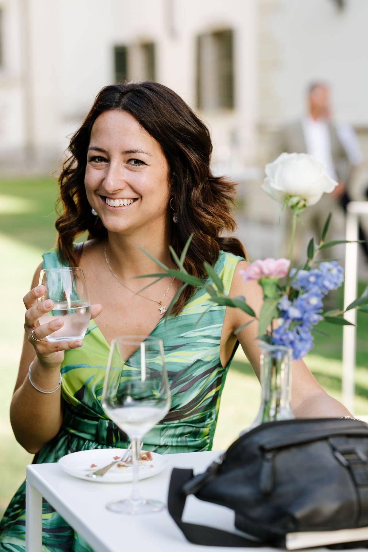 Attendee in green dress enjoying wine at outdoor event with floral centerpiece on table.