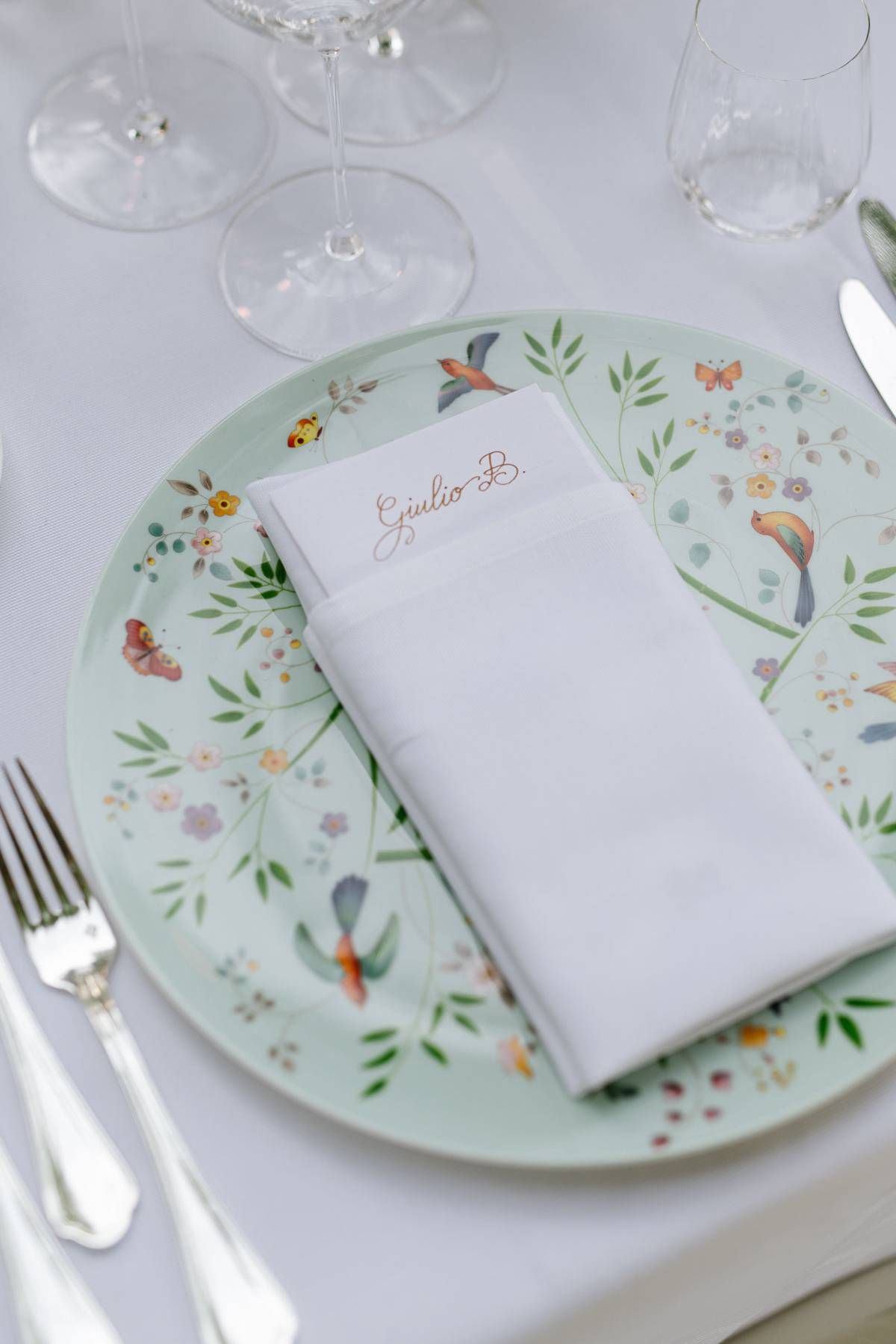 Wedding place setting with floral patterned plate and white napkin on tablecloth.