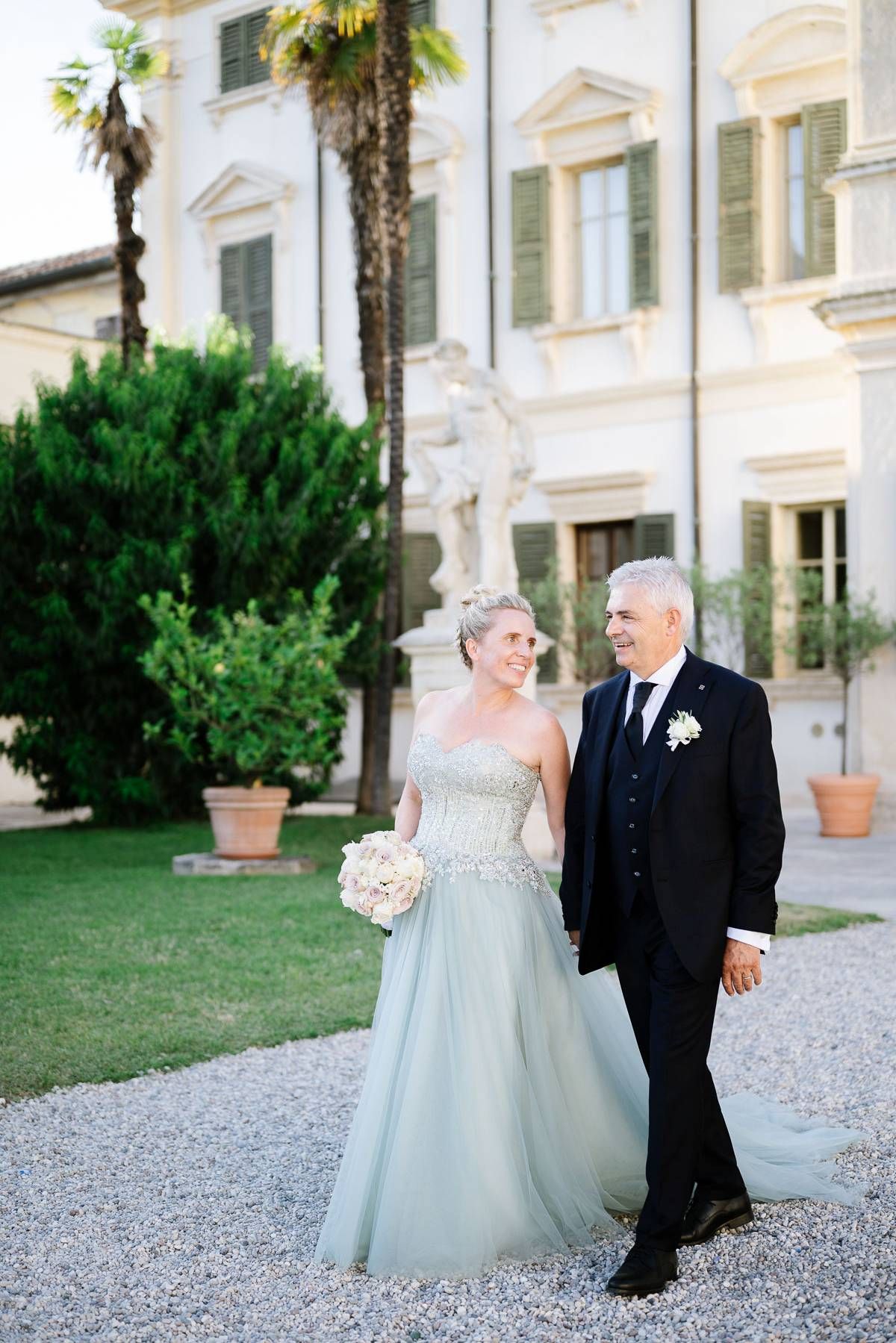 Couple in formal attire walking outside elegant Italian villa with palm trees.