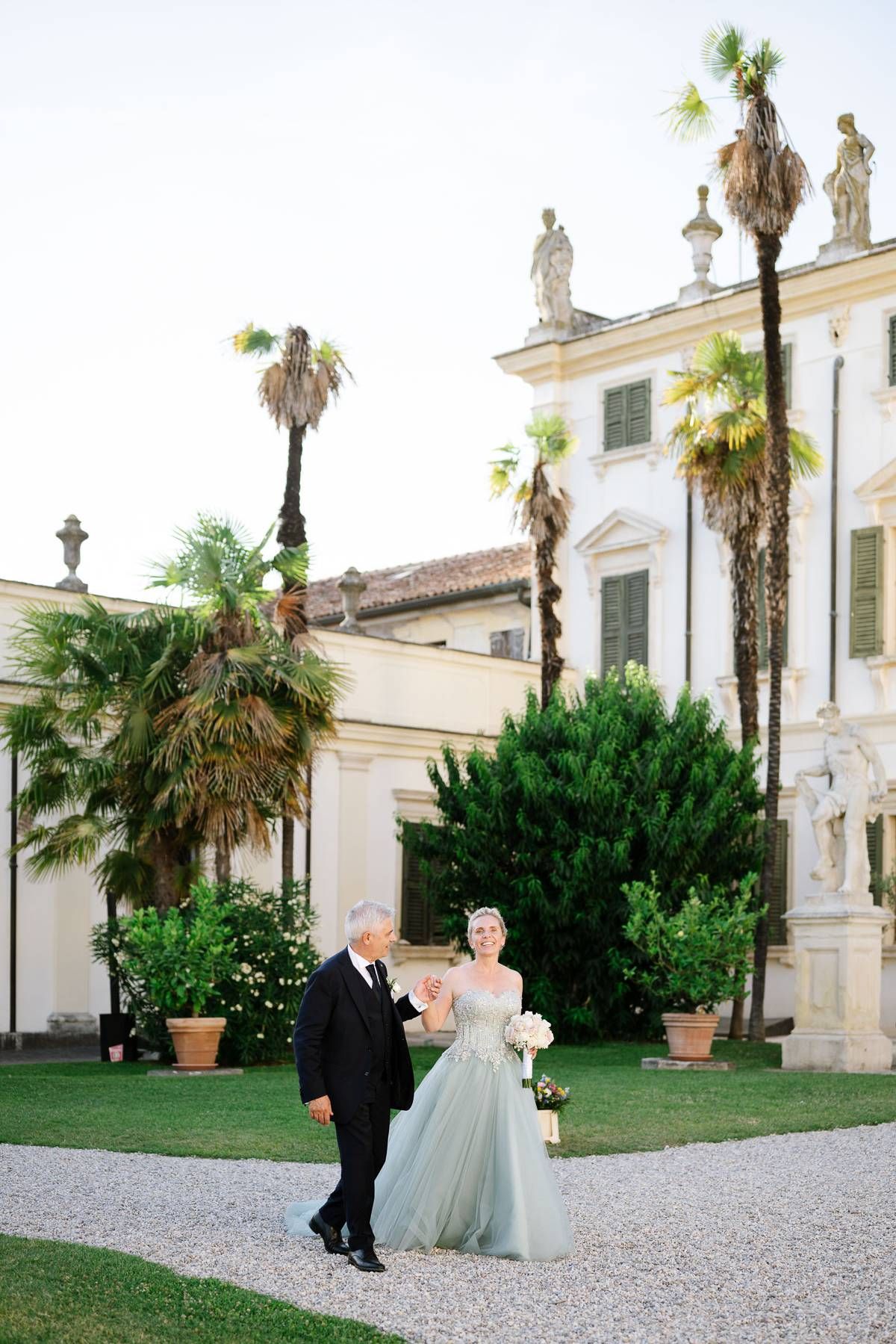 A couple walks on a gravel path in front of an elegant white mansion with palm trees and ornate architectural details.