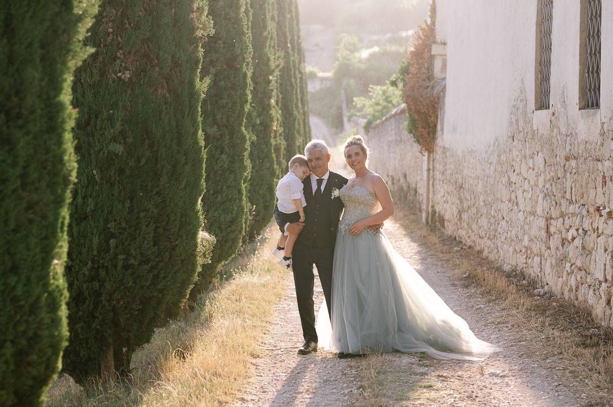 A romantic wedding moment captured between cypress trees along a sunlit path.