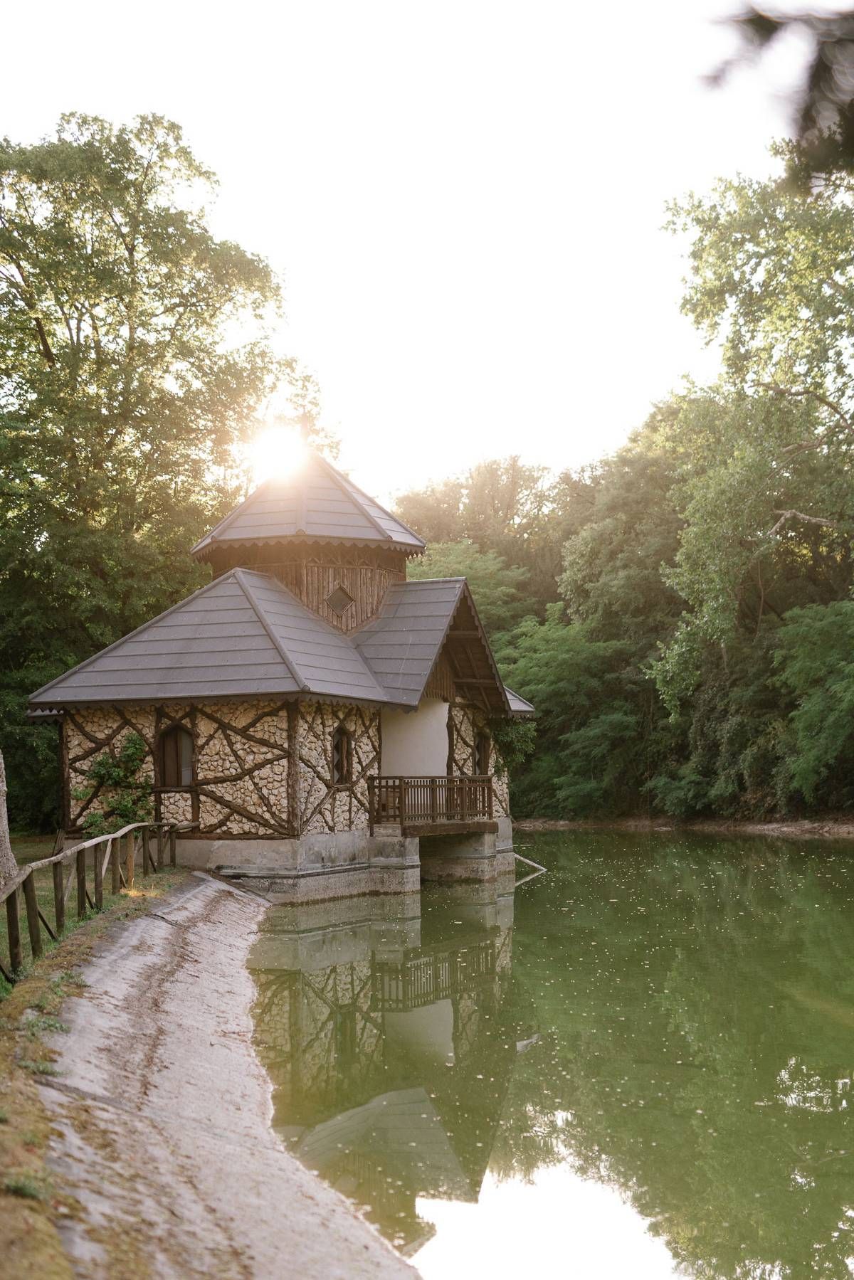 Rustic wooden gazebo on a pond reflecting sunset light through surrounding trees.