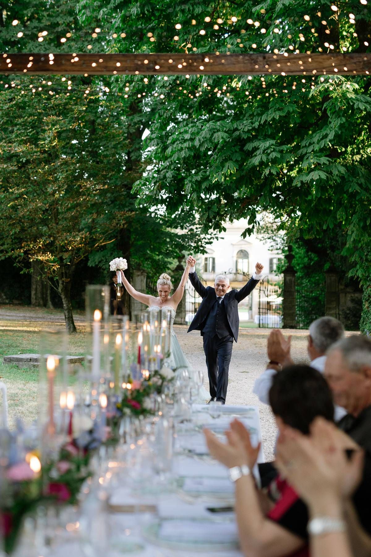 Joyful wedding celebration with guests clapping at a long candlelit outdoor table.