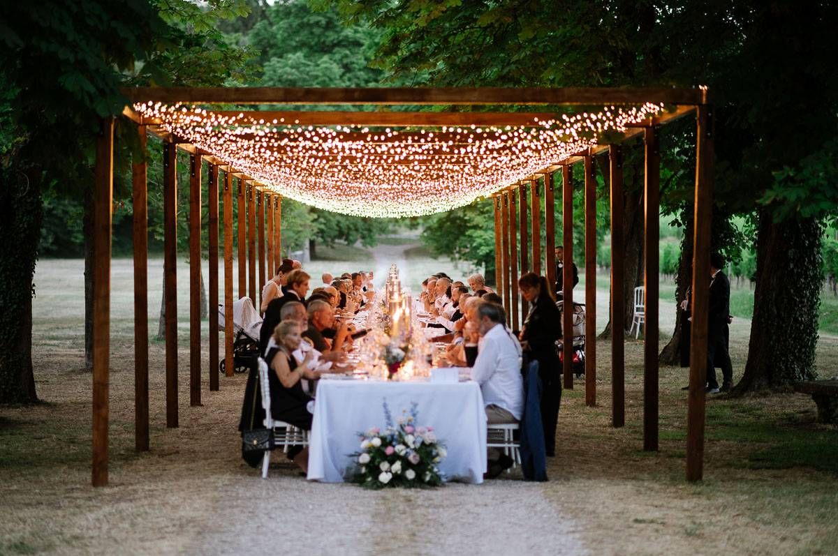 Elegant outdoor dinner party under illuminated wooden pergola with long white dining table.