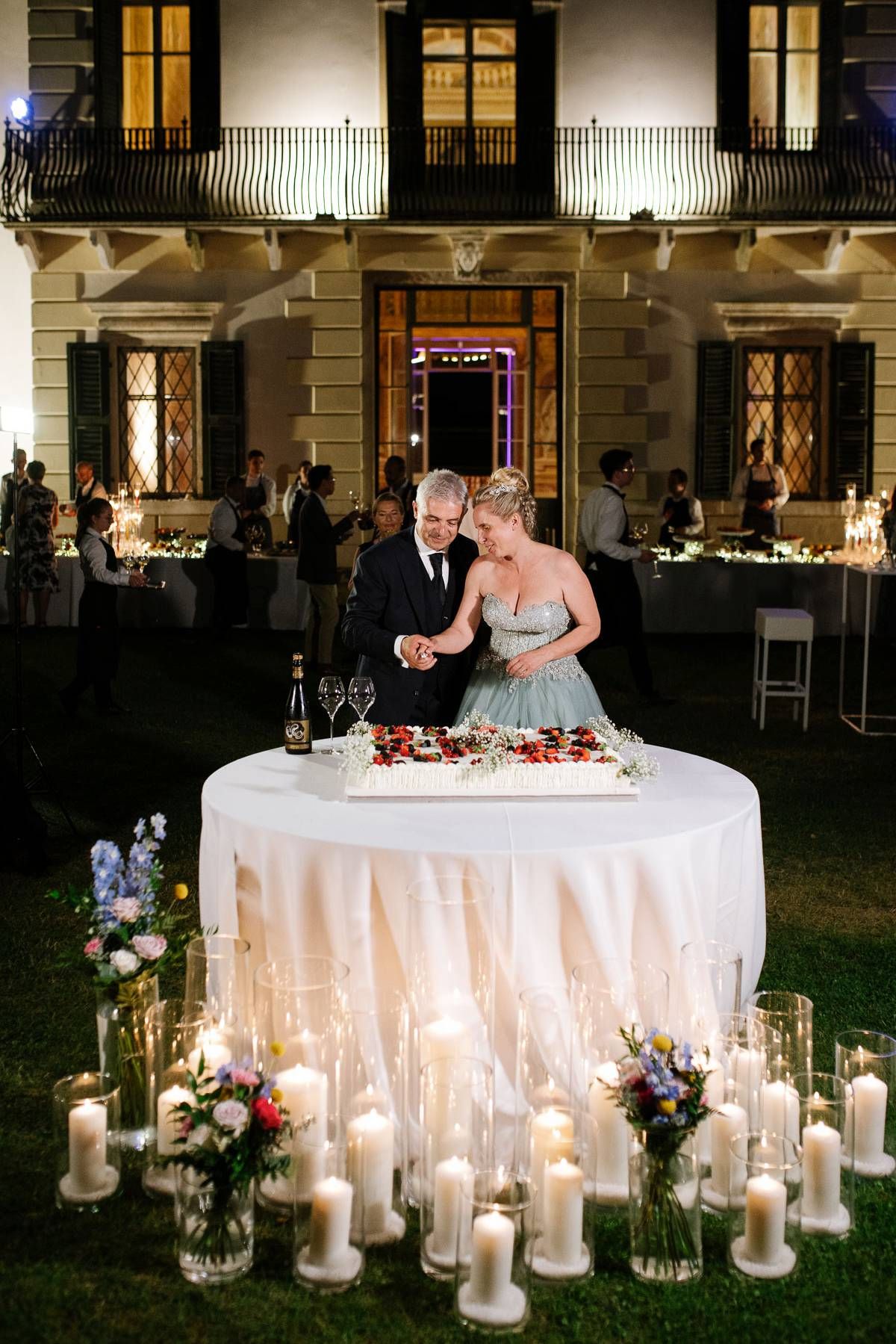Romantic wedding cake table surrounded by glowing candles against elegant building facade at night.
