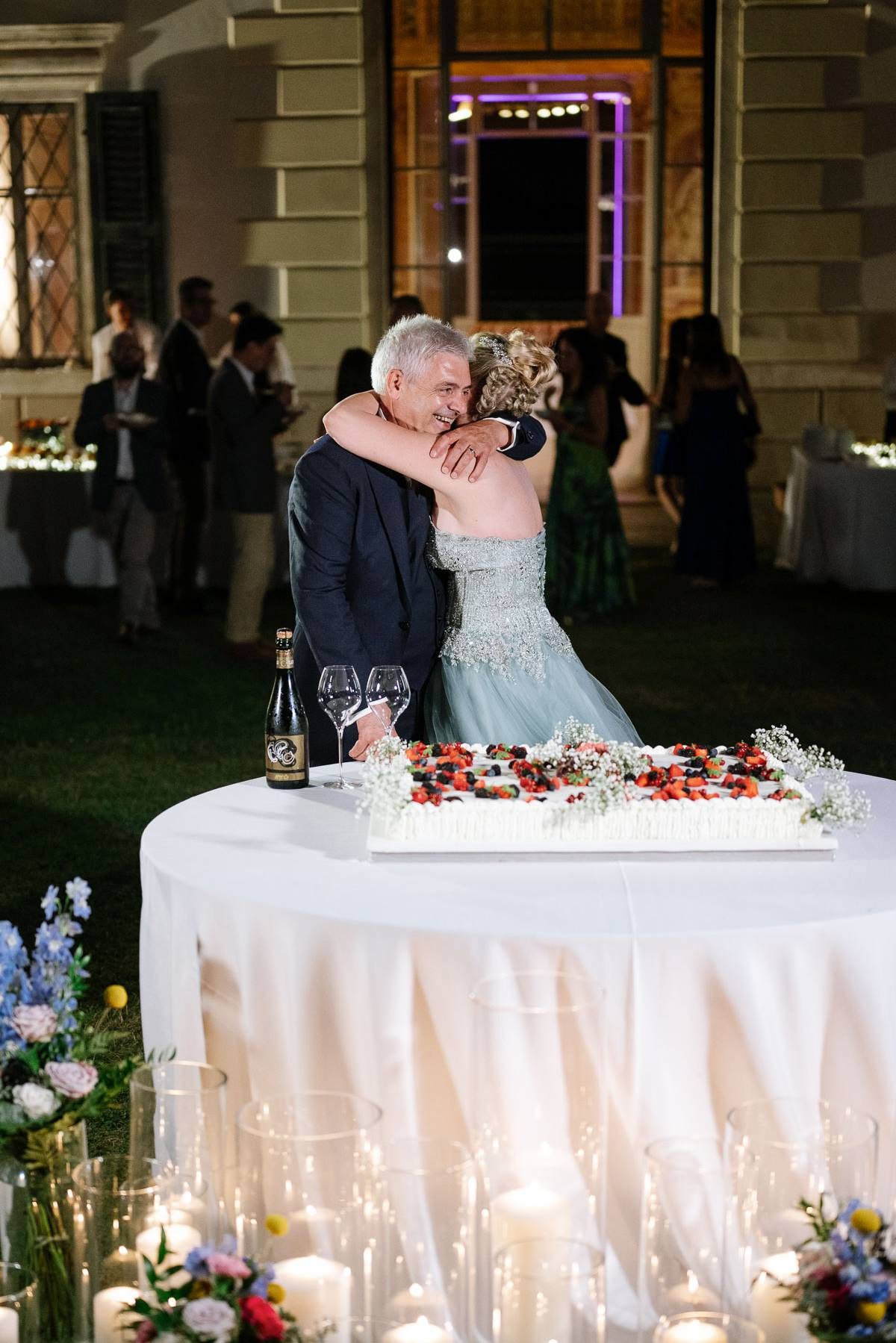 Newlyweds embrace tenderly behind their wedding cake decorated with red berries.