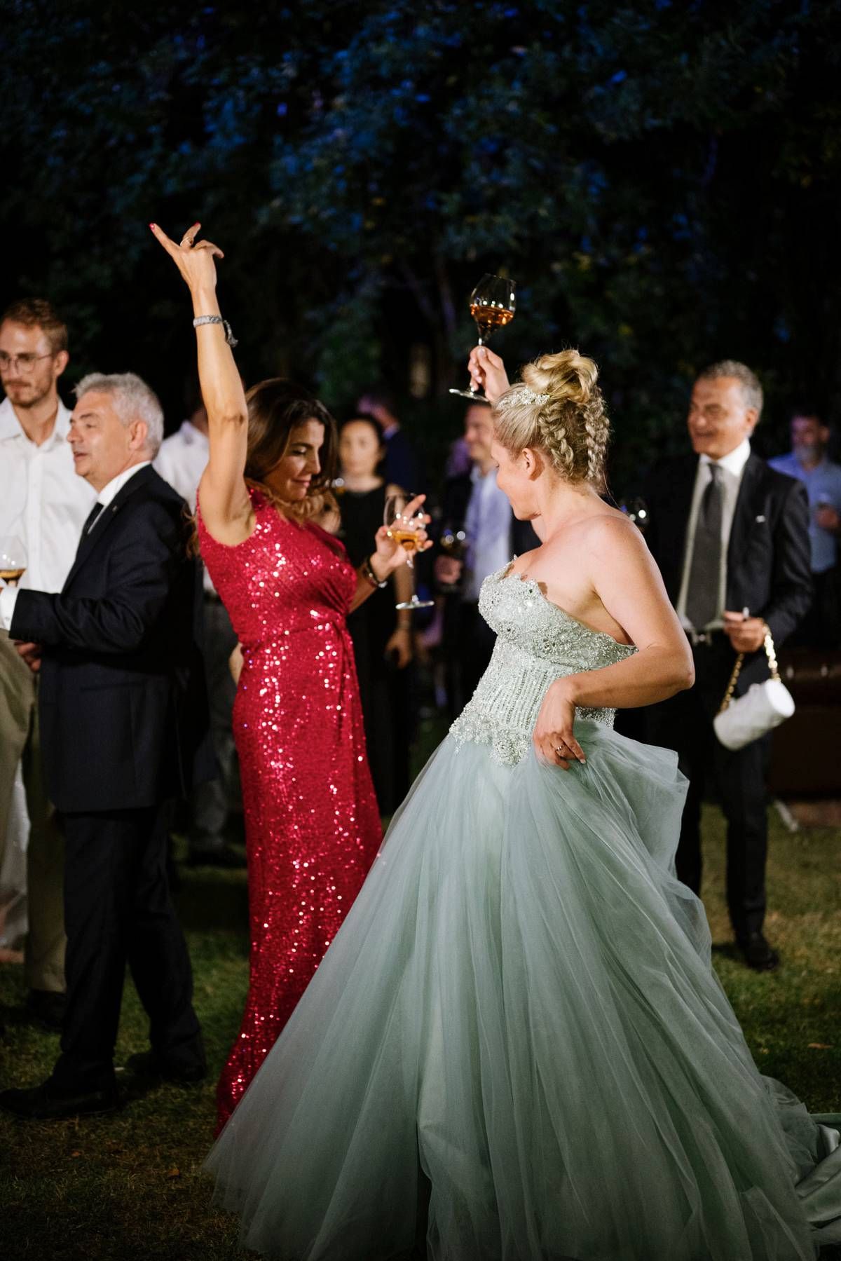 Two guests in evening wear dance enthusiastically at an outdoor reception with trees in background.
