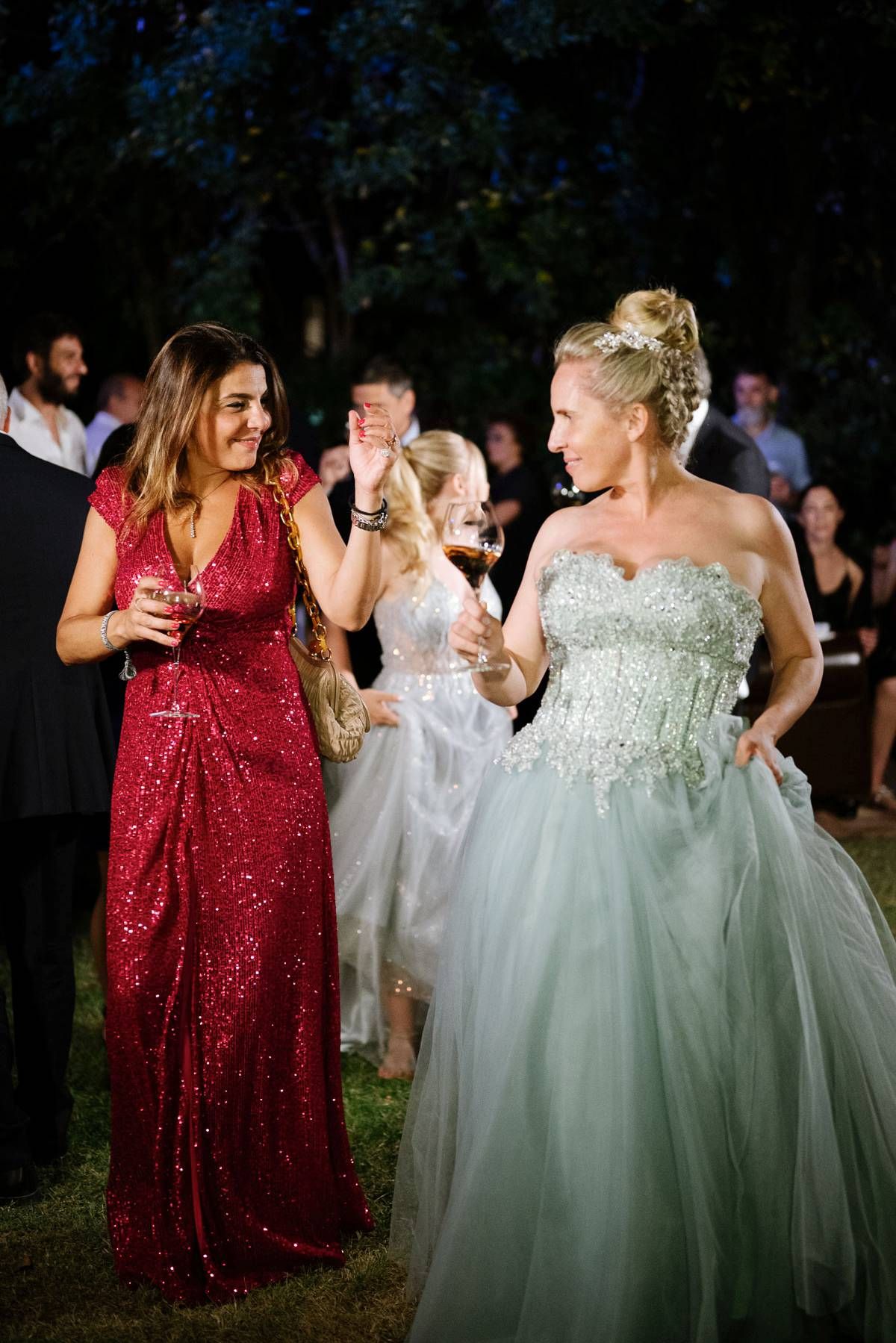 Two guests in formal attire, one in red sequins and one in a mint ballgown, enjoying drinks at an outdoor reception.
