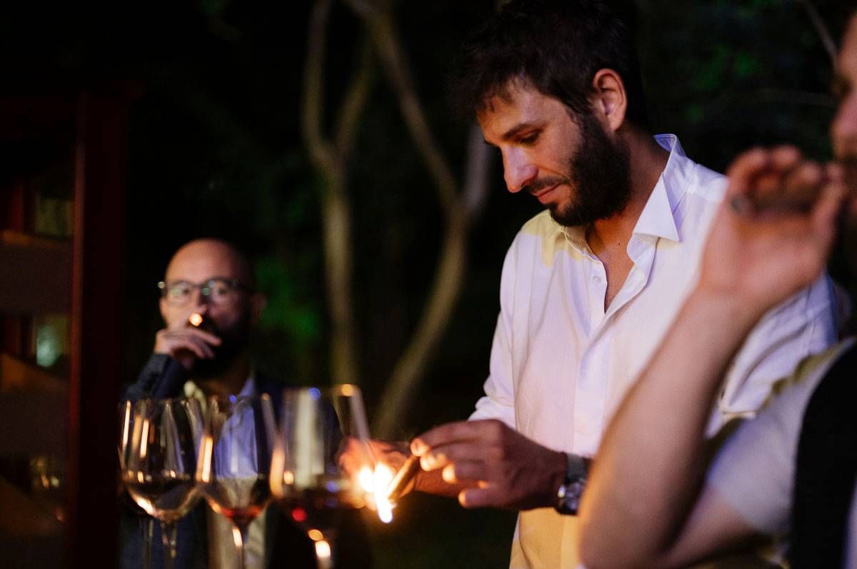 Guest lighting a sparkler at evening wedding reception with wine glasses in foreground.