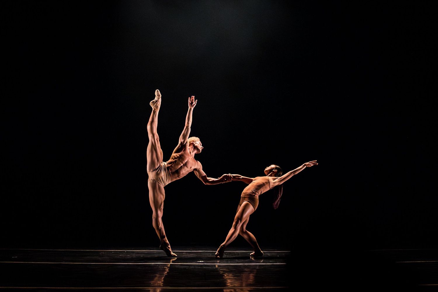 Two dancers perform synchronized contemporary movements against dark backdrop.