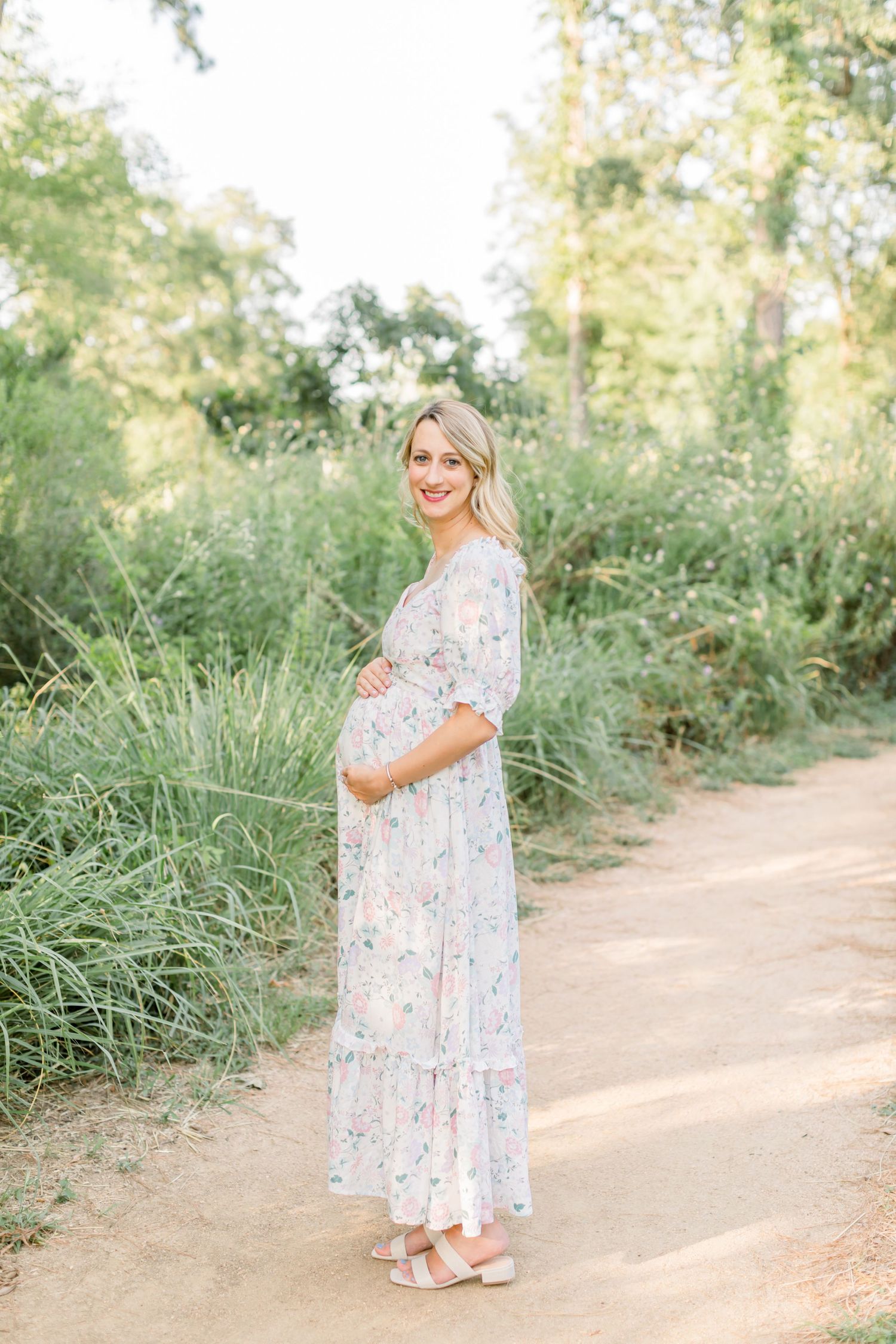 A radiant mother-to-be in a floral dress stands on a sunlit forest path.