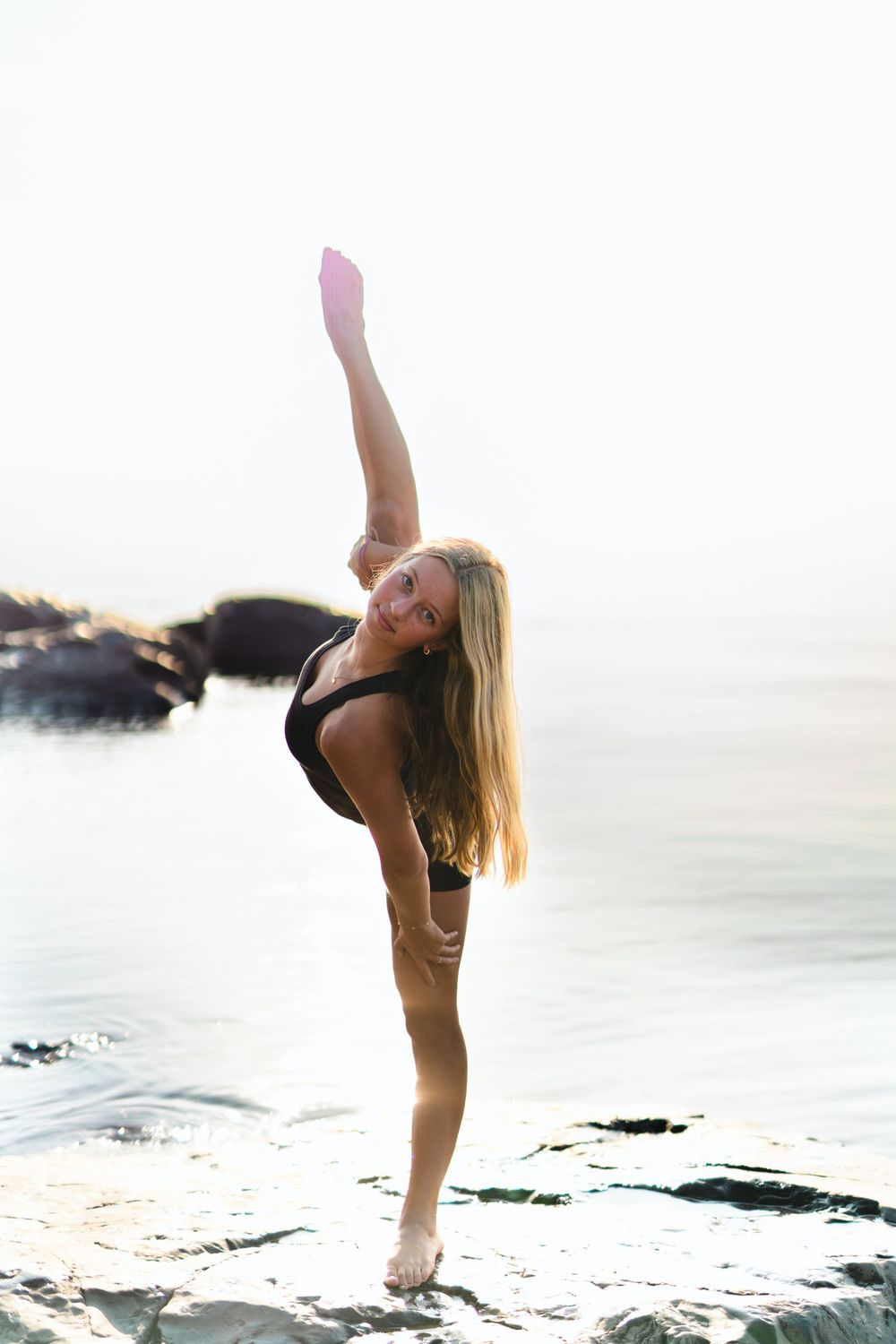 Dancer performing high kick pose on rocky beach at sunset.