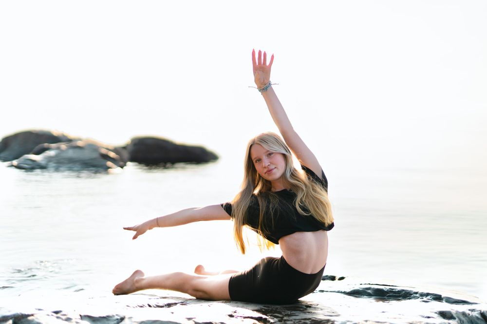 Dancer in black outfit performing graceful pose on rocky beach.