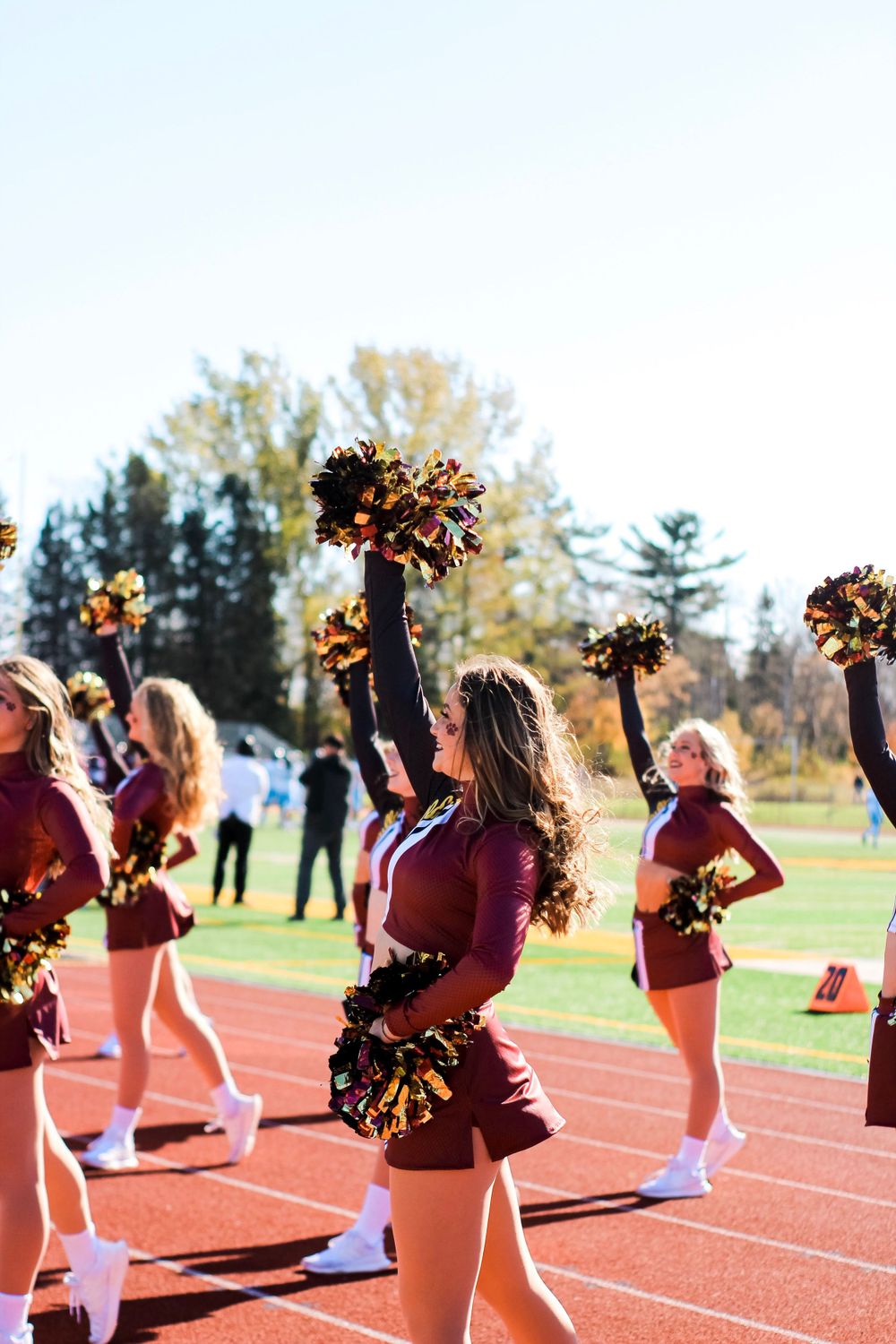 Cheerleaders in maroon uniforms performing with pom poms on an outdoor track field during a sunny day.