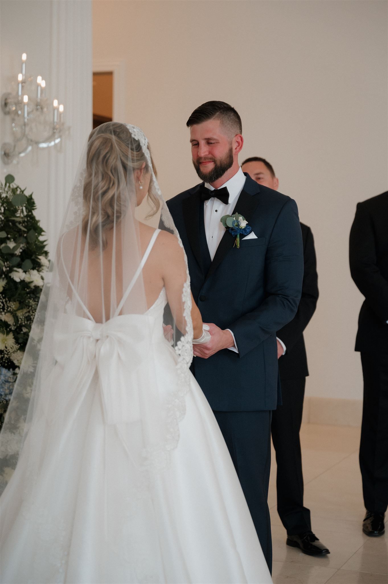 A wedding ceremony moment in an elegant white room with chandelier and floral decor.