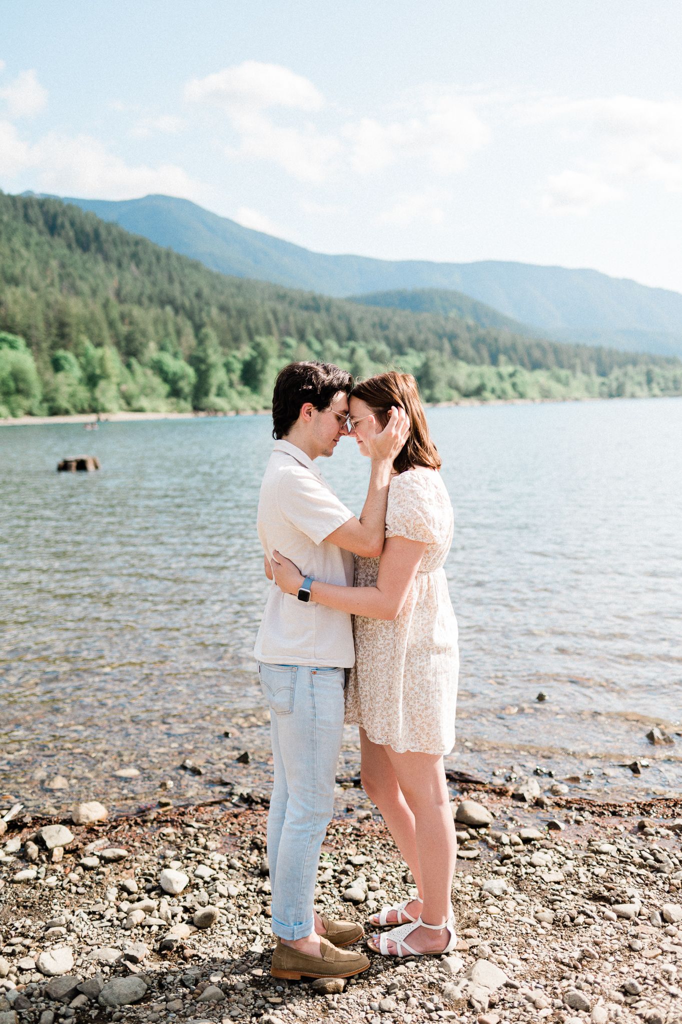 A couple stands together forehead to forehead by a scenic mountain lake with evergreen forests in the background.