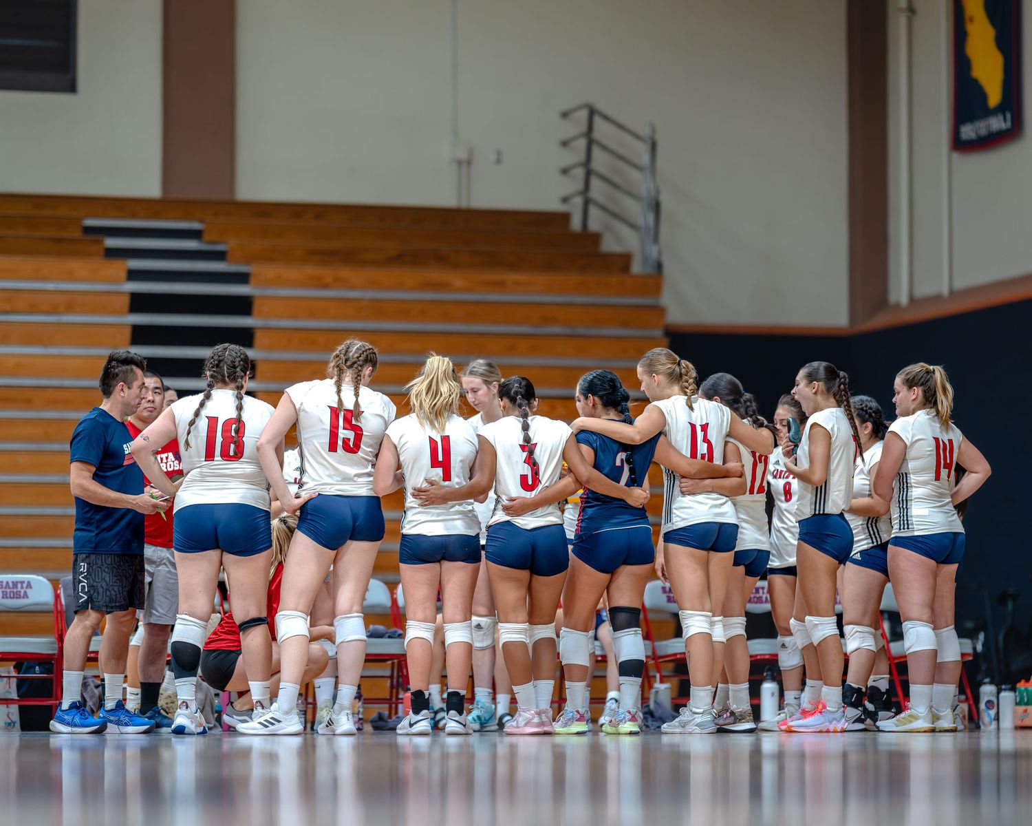 Women's volleyball team huddles with coach during game strategy discussion in indoor gymnasium.