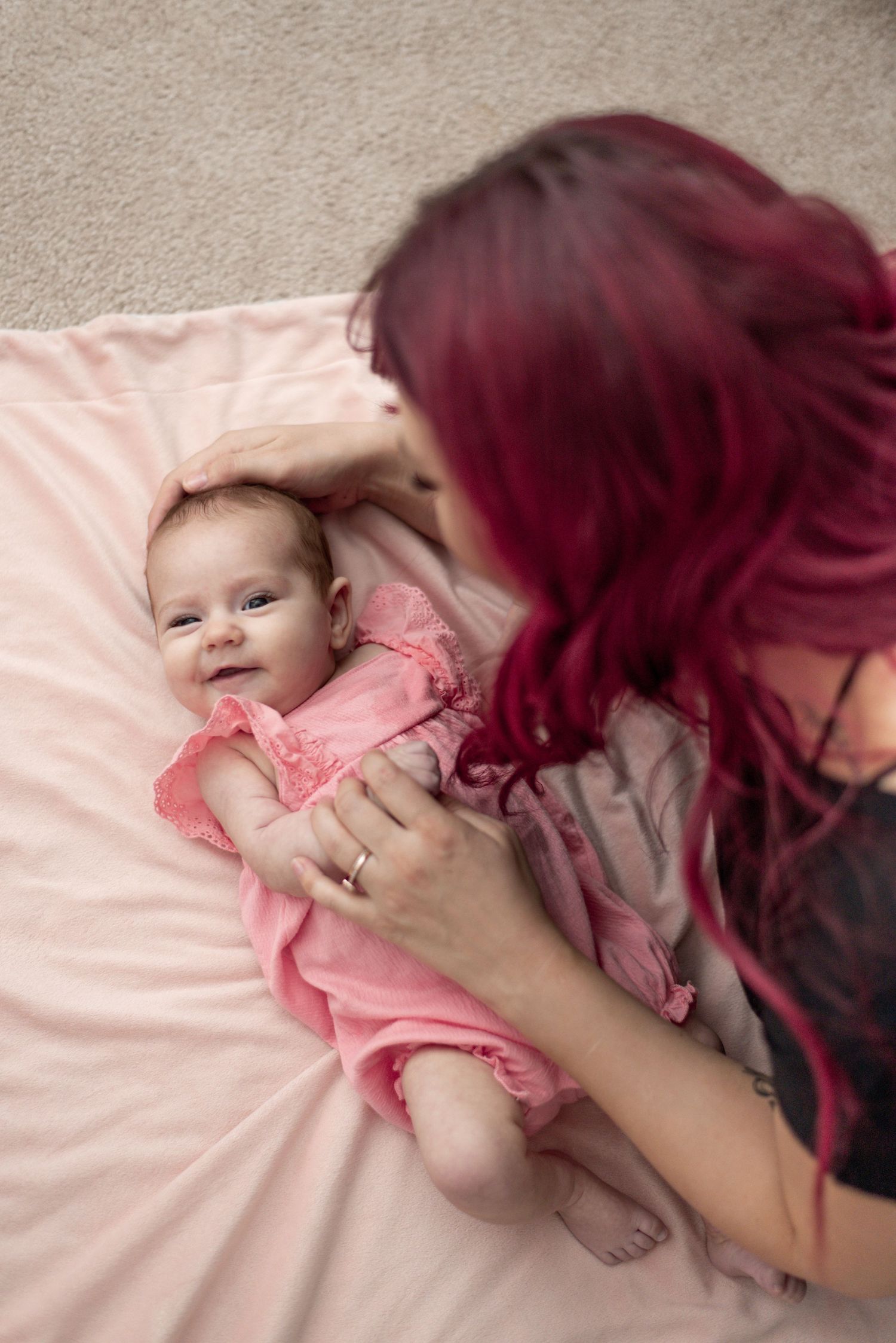 A baby in pink clothing lies on a soft bed while being gently touched.