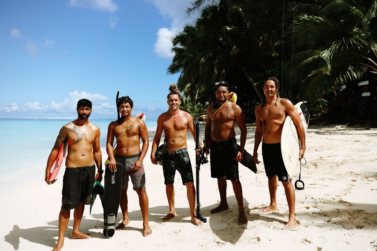 Group of snorkelers with gear pose together on tropical beach with palm trees and blue water.