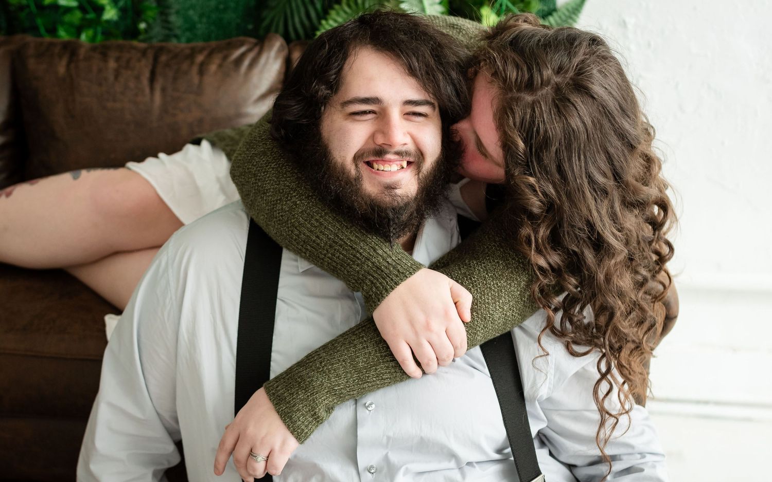 Man sitting in front of couch while his wife who is laying on the couch wraps her arms around him and whispers to him
